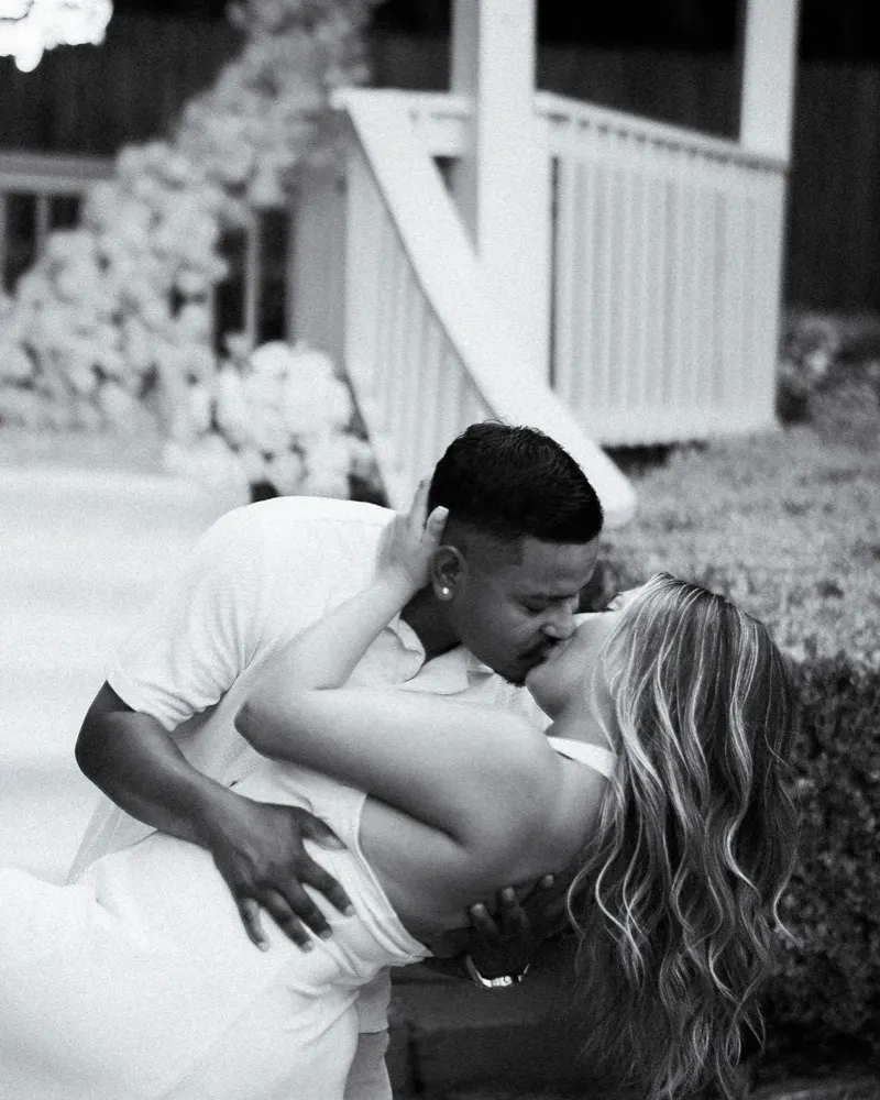 Black and white photo of a man dipping and kissing his fiancée in a garden with a white floral arch